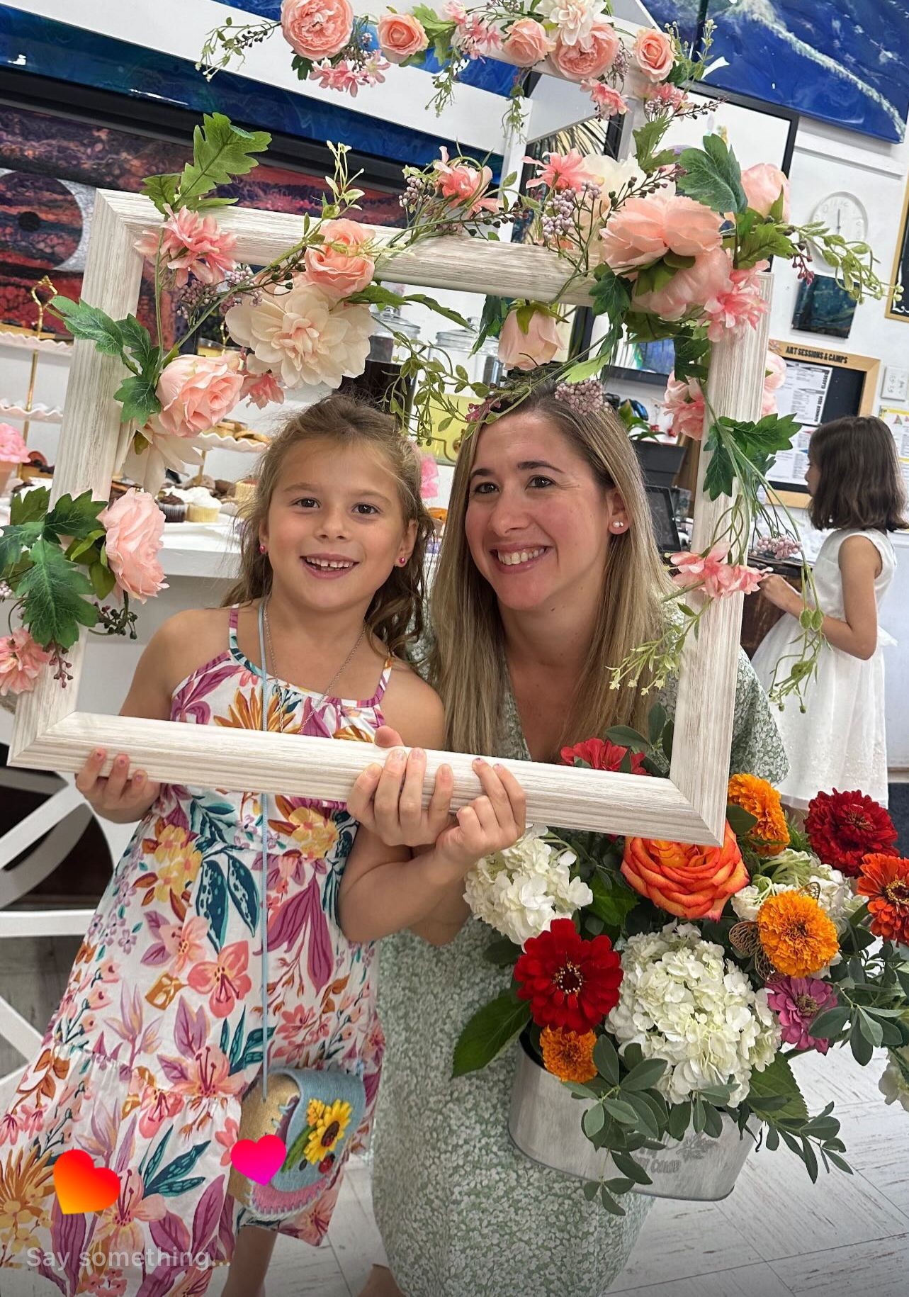 Smiling woman and girl holding a floral frame, surrounded by colorful flowers indoors.