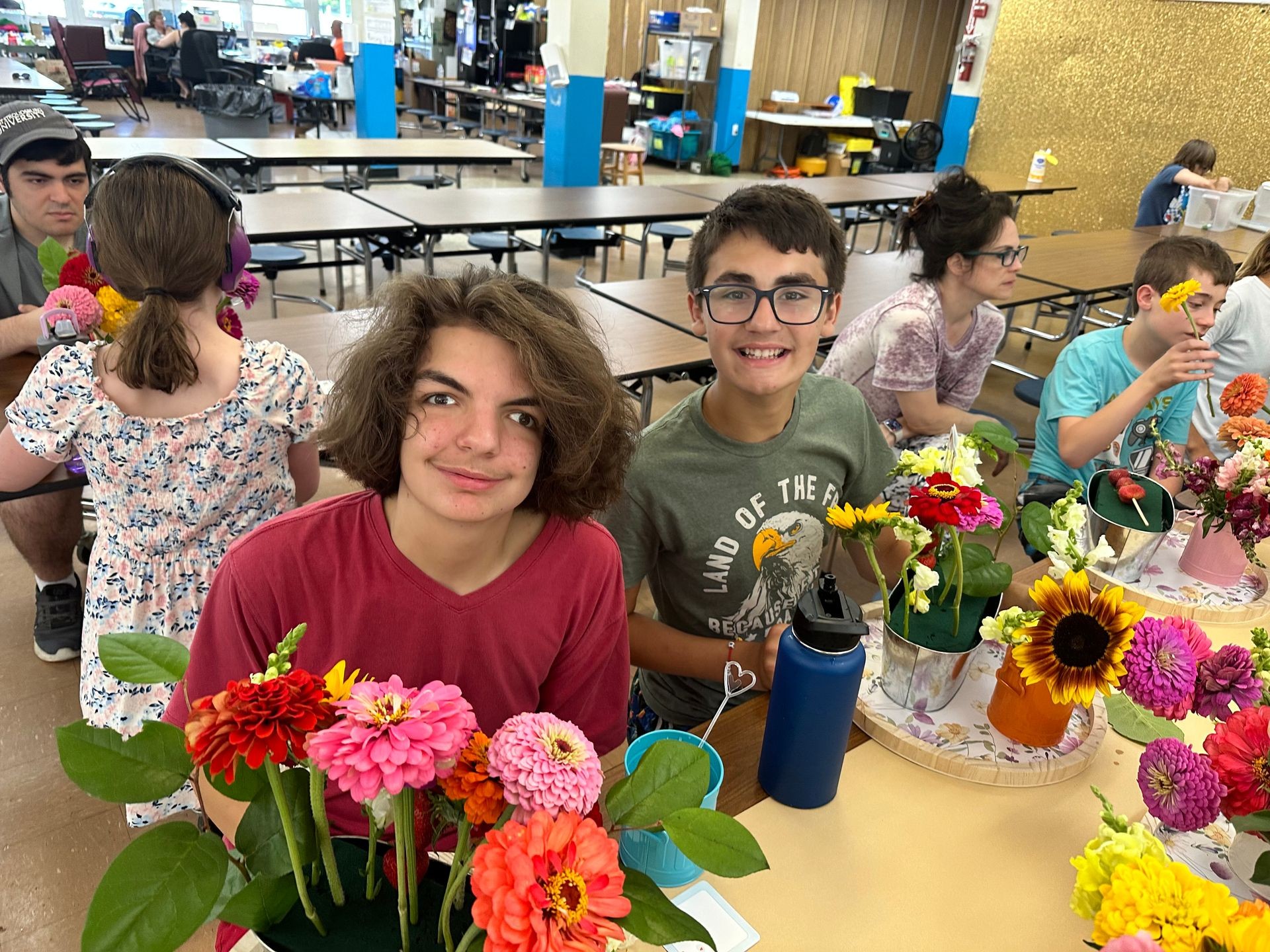 Children sitting at a table in a classroom, surrounded by colorful arrangements of flowers and plants.